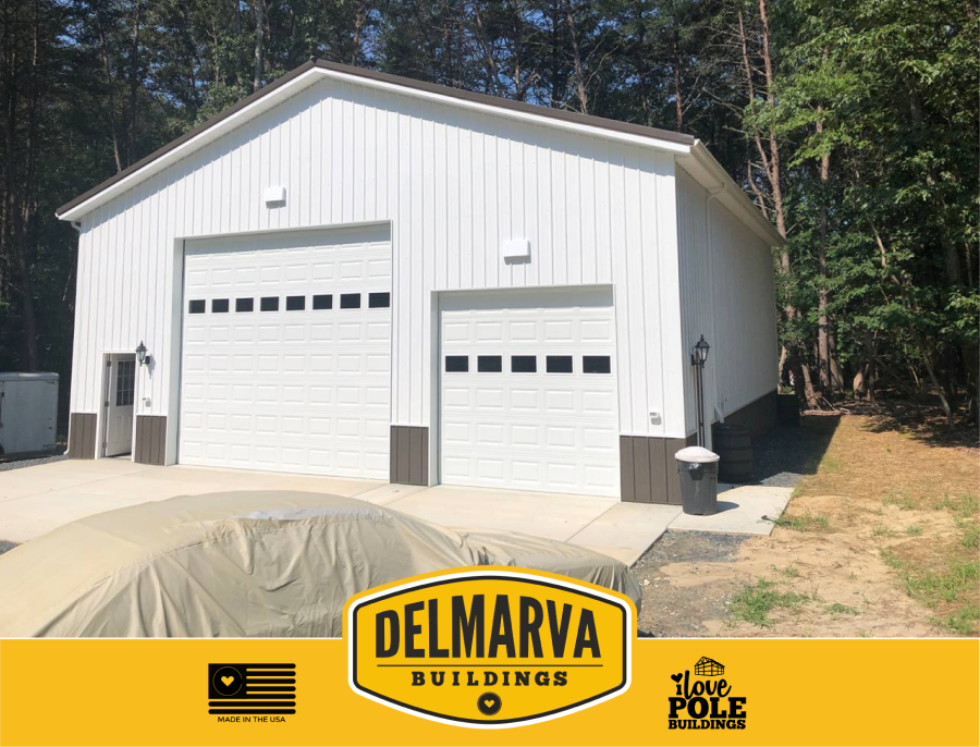 White metal garage building with two white garage doors and brown trim, surrounded by pine trees.