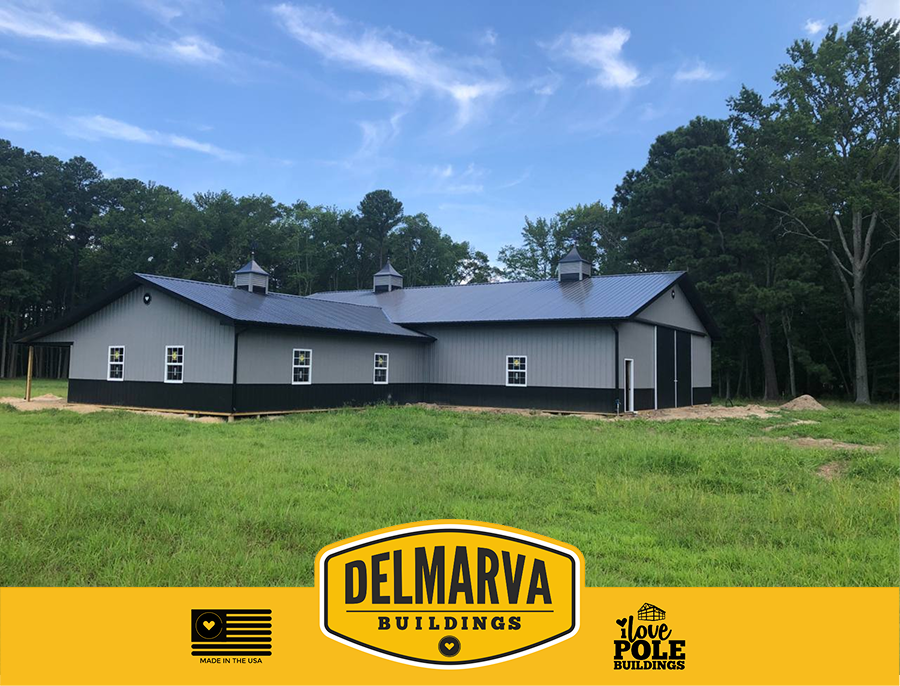 Gray pole barn with black trim, metal roof, and multiple windows surrounded by trees .