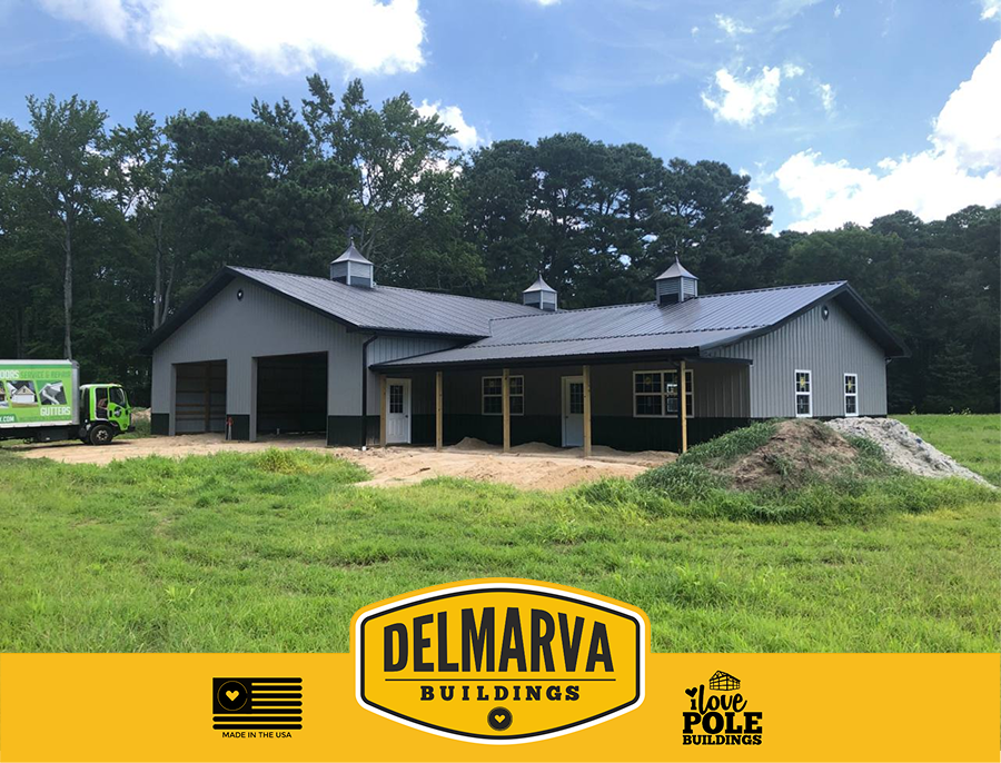 Gray pole barn with black trim, covered porch, and metal roof surrounded by trees.