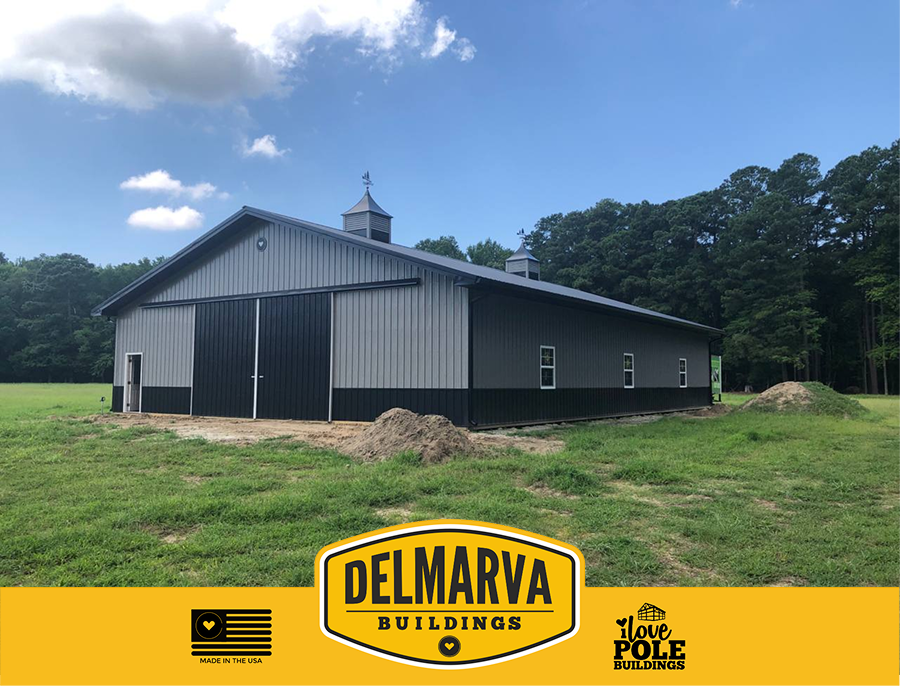 Black and gray pole barn with cupolas and large doors surrounded by trees.