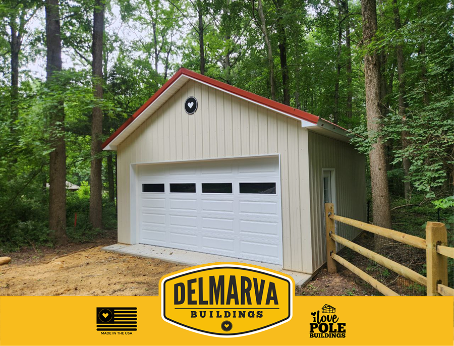 Cream pole barn with red roof and white garage door surrounded by trees.