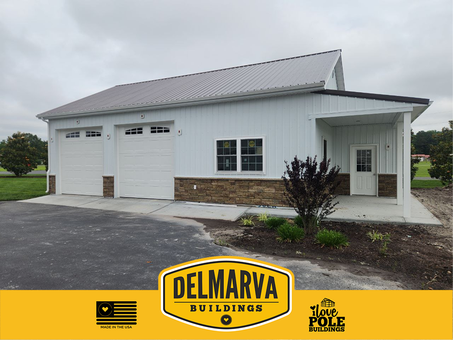 White pole barn house with two white garage doors, stone base, and covered porch.