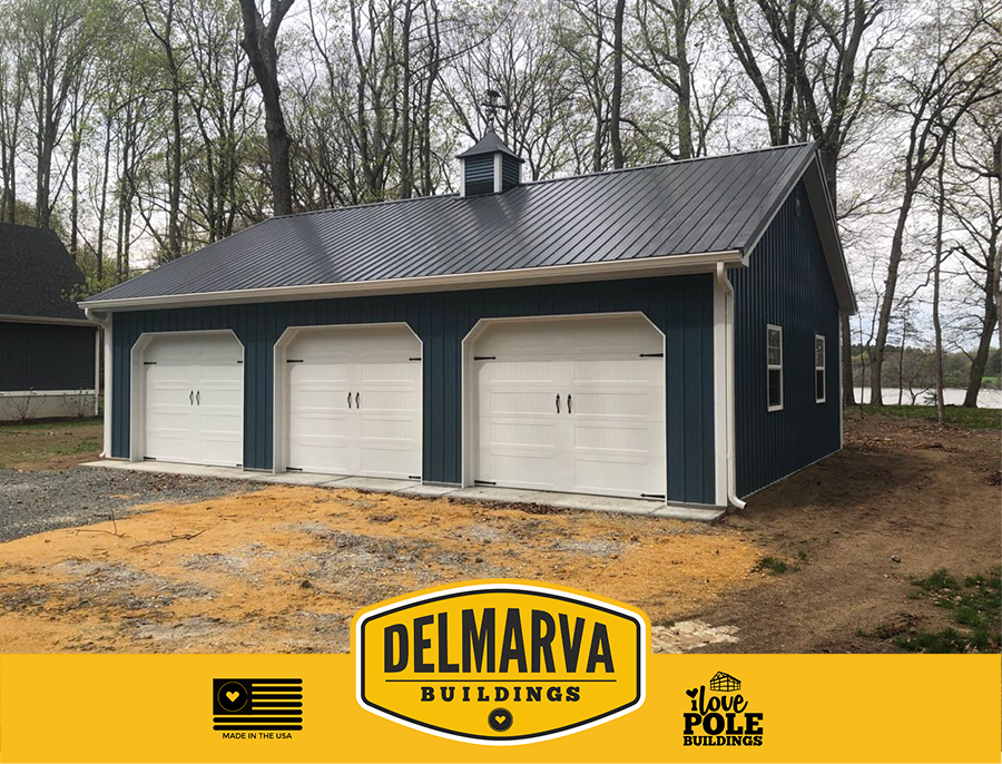 Dark blue pole barn with three white garage doors, cupola, and white trim by Delmarva Buildings.