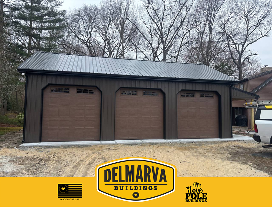 Dark gray pole barn with three brown garage doors and metal roof by Delmarva Buildings.